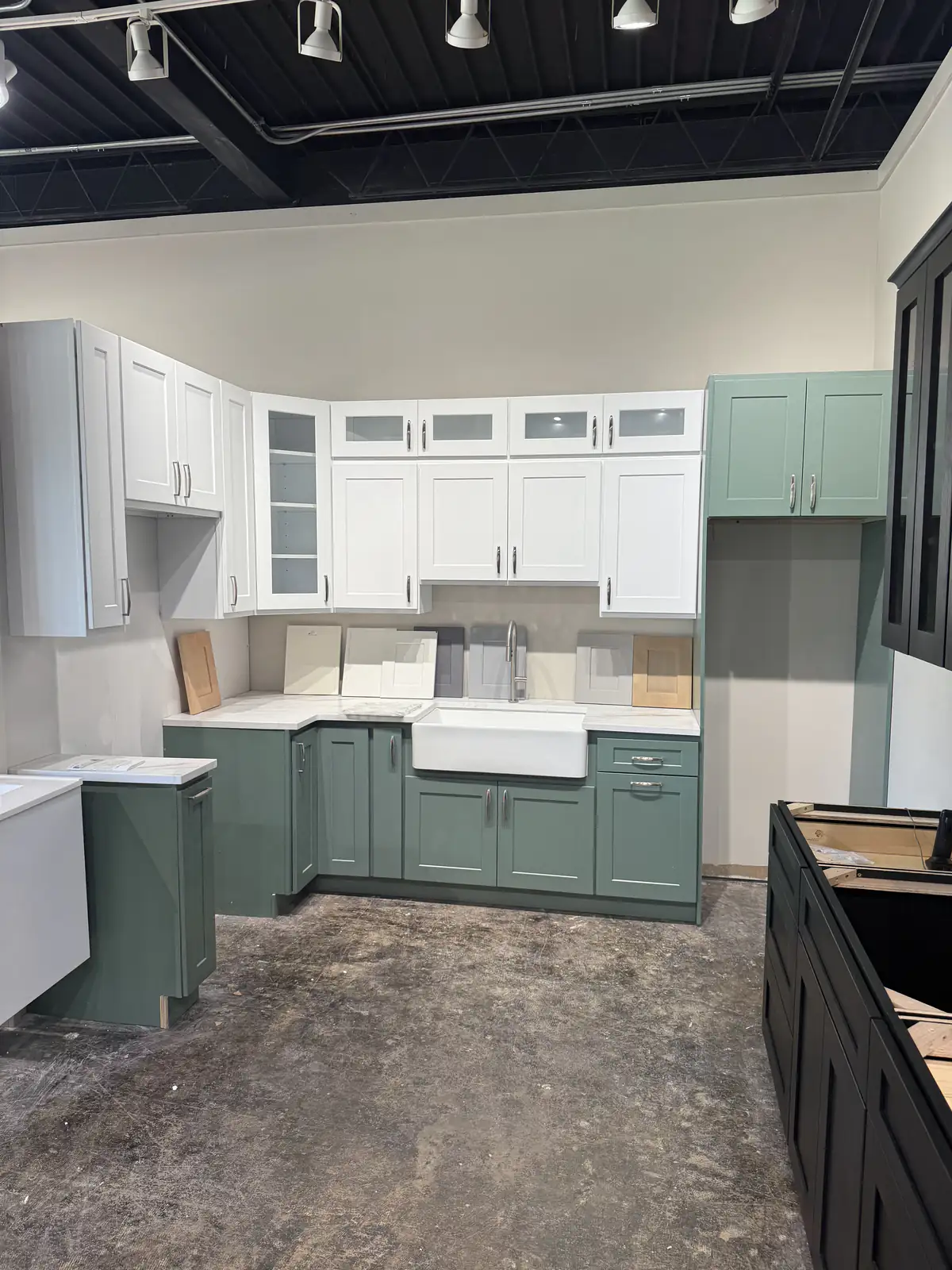 Kitchen displays in the Lexington showroom: white shaker uppers over sage green base cabinets with a farmhouse sink and door samples on the counter