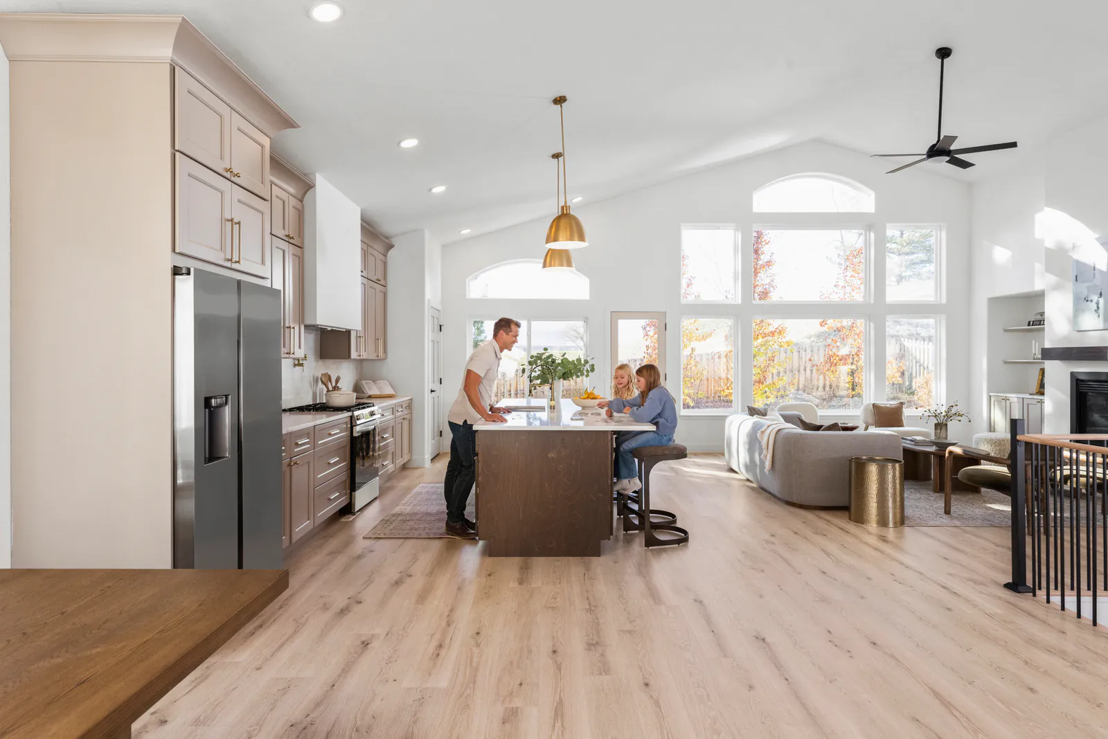 A family at a kitchen island in an open kitchen with taupe cabinets, brass pendant lights and tall windows
